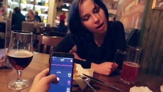 Красотка с темными волосами сидит в кафе с другом, который управляет со смартфона игрушкой в ее письке