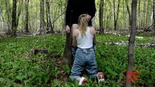Девушка в джинсах и в белой майке встает на колени возле дерева и сосет партнеру